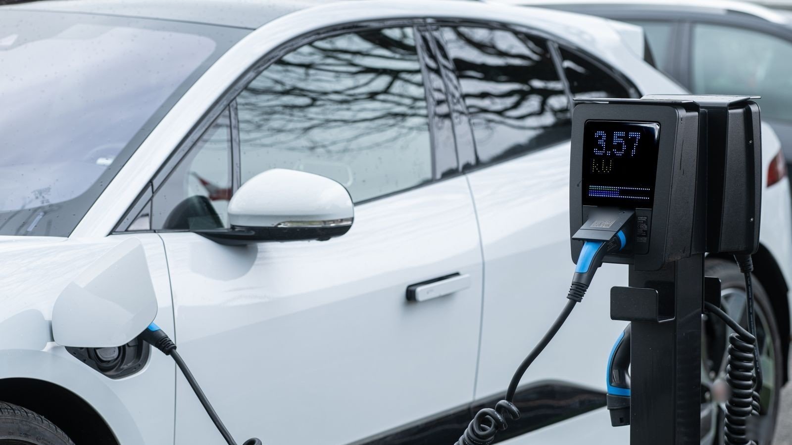 Weißes Elektroauto an einer Ladesäule, der Ladestecker steckt in der Buchse am vorderen Kotflügel, das Display der Säule zeigt die Ladeleistung