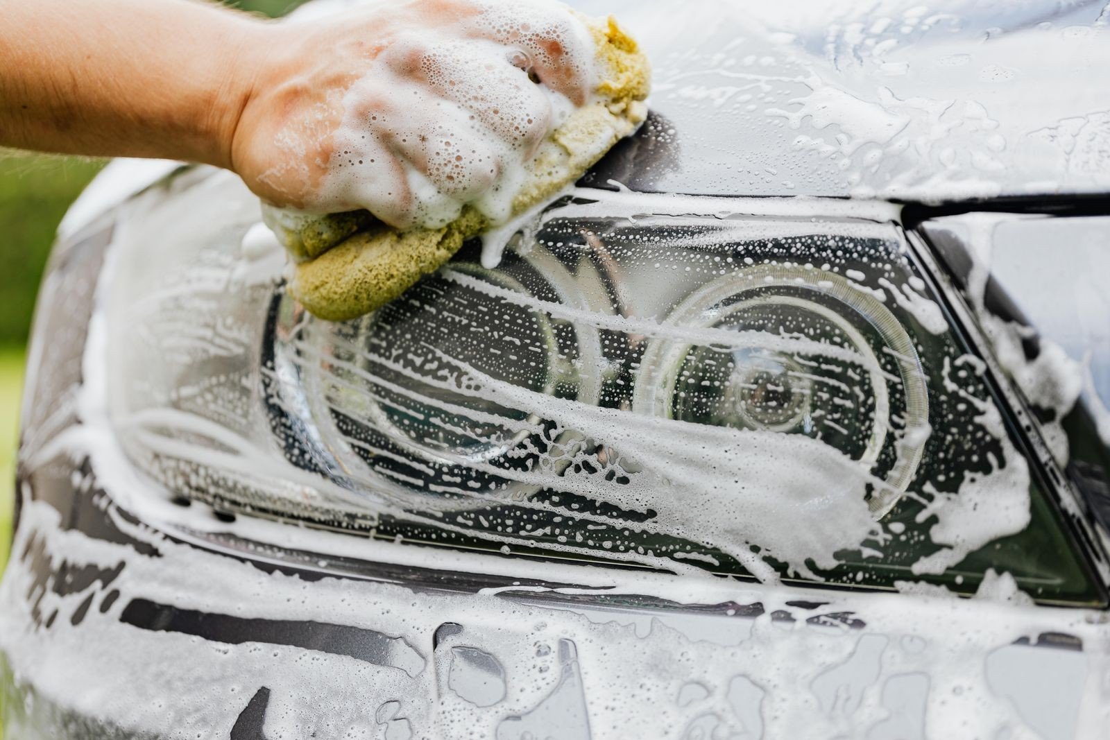Hand wäscht mit einem gelben Schwamm und dichtem Schaum den Scheinwerfer eines hellen Autos