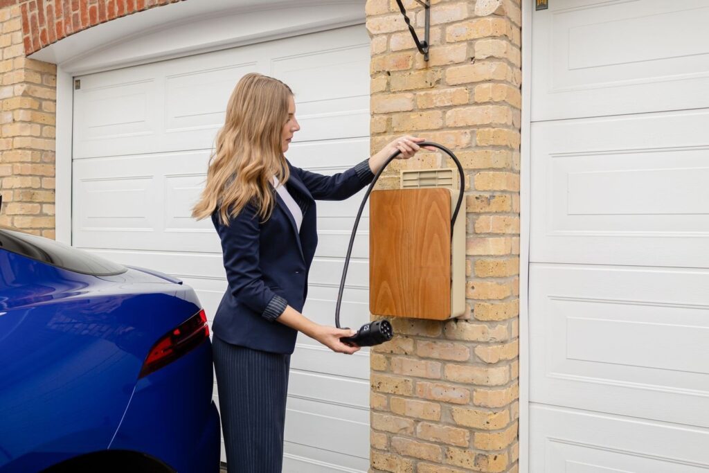 Frau steckt den Ladestecker einer holzverkleideten Wallbox an der Klinkerwand neben dem Garagentor ein, davor ein blaues Auto